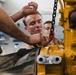 Several individuals, appearing to be military personnel or trainees in a workshop setting, are working on a large yellow industrial or military engine component. A chain hoist with a red hook is being used to assist with the mechanical work. The scene suggests a hands-on maintenance training exercise.