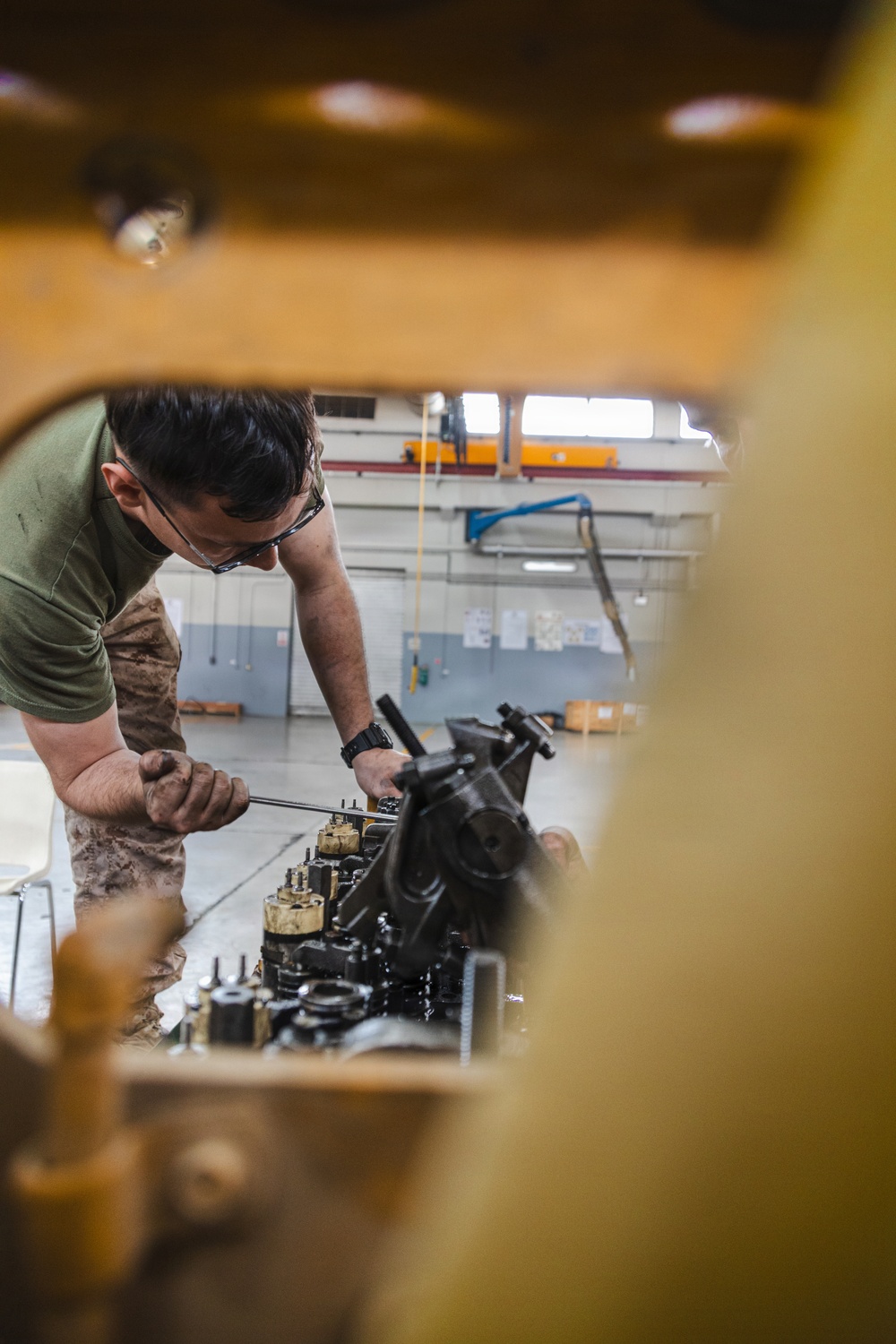 A mechanic wearing a green t-shirt and camouflage pants is working on a large engine or mechanical assembly inside an industrial maintenance bay. The photo is taken through an opening in what appears to be yellow heavy equipment, framing the worker. The setting appears to be a military or industrial vehicle maintenance facility.