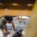 A mechanic wearing a green t-shirt and camouflage pants is working on a large engine or mechanical assembly inside an industrial maintenance bay. The photo is taken through an opening in what appears to be yellow heavy equipment, framing the worker. The setting appears to be a military or industrial vehicle maintenance facility.