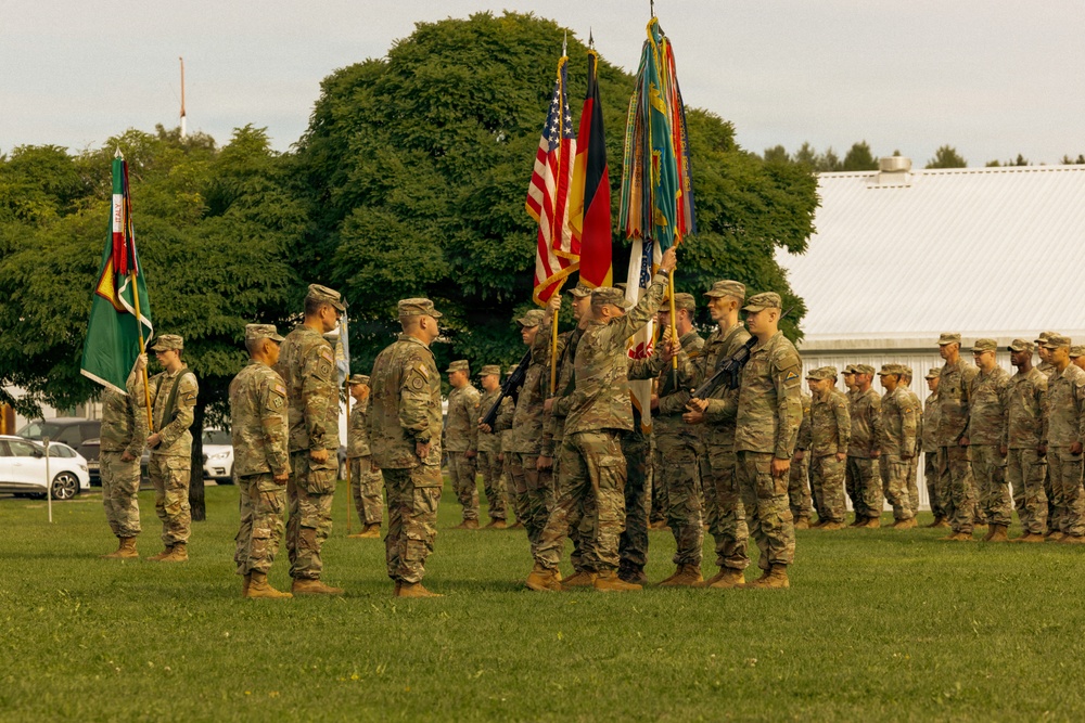 A formal military ceremony is taking place on a grass parade field, likely a change of command or unit activation/deactivation ceremony. Soldiers in US Army OCP (Operational Camouflage Pattern) uniforms are assembled in formation while a color guard presents multiple flags including the US flag and German flag. The setting appears to be a military installation in Germany based on the visible flags and architecture.