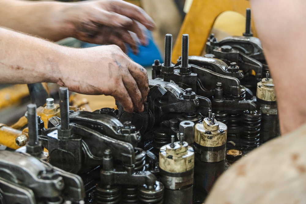 Two mechanics are working on a large diesel engine, likely a Caterpillar (CAT) engine, with their hands visibly dirty with grease and oil. The image shows a close-up of the engine's valve train or cylinder head assembly with multiple components exposed. The work appears to be taking place in a workshop or garage environment.