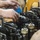 Two mechanics are working on a large diesel engine, likely a Caterpillar (CAT) engine, with their hands visibly dirty with grease and oil. The image shows a close-up of the engine's valve train or cylinder head assembly with multiple components exposed. The work appears to be taking place in a workshop or garage environment.