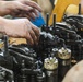 Two mechanics are working on a large diesel engine, likely a Caterpillar (CAT) engine, with their hands visibly dirty with grease and oil. The image shows a close-up of the engine's valve train or cylinder head assembly with multiple components exposed. The work appears to be taking place in a workshop or garage environment.