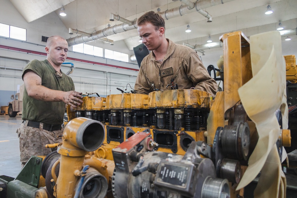 Two military personnel are working on a large yellow diesel engine in what appears to be a military maintenance bay or motor pool facility. One individual is wearing a green t-shirt with camouflage trousers, while the other is wearing tan/khaki coveralls, suggesting different roles or units. The engine, painted yellow and consistent with heavy equipment or generator machinery, is partially disassembled on a workbench.