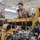 Two military personnel are working on a large yellow diesel engine in what appears to be a military maintenance bay or motor pool facility. One individual is wearing a green t-shirt with camouflage trousers, while the other is wearing tan/khaki coveralls, suggesting different roles or units. The engine, painted yellow and consistent with heavy equipment or generator machinery, is partially disassembled on a workbench.