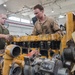 Two military personnel are working on a large yellow diesel engine in what appears to be a military maintenance bay or motor pool facility. One individual is wearing a green t-shirt with camouflage trousers, while the other is wearing tan/khaki coveralls, suggesting different roles or units. The engine, painted yellow and consistent with heavy equipment or generator machinery, is partially disassembled on a workbench.