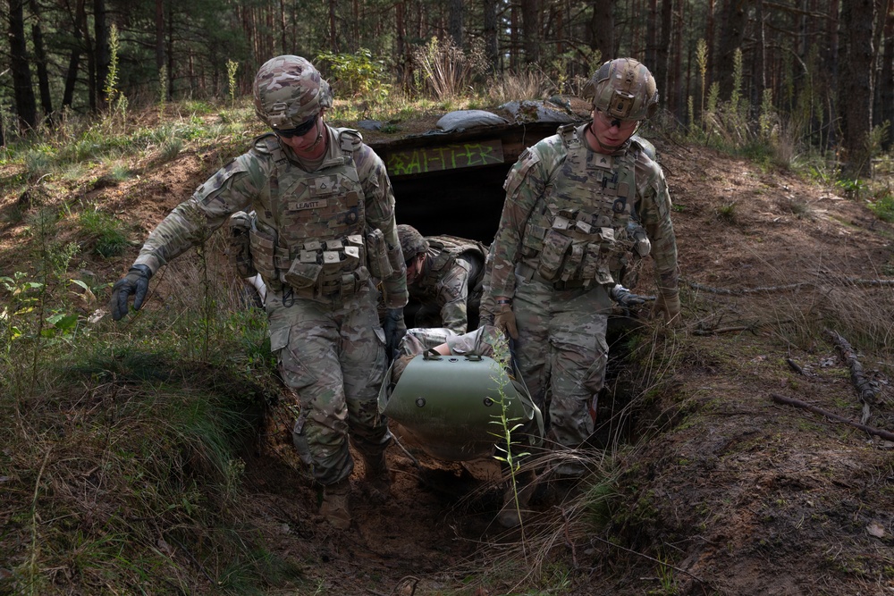 Two U.S. Army soldiers in MultiCam (OCP) uniforms are extracting a simulated casualty on a litter from a field bunker or dugout structure in a forested training area. The soldier on the left is identified by a name tape reading 'LEAVITT.' This appears to be a tactical combat casualty care (TCCC) or medevac training exercise.