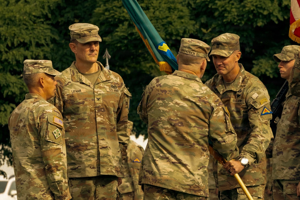 A U.S. Army military ceremony is taking place outdoors, likely a change of command or unit activation event. Several soldiers in MultiCam/OCP camouflage uniforms are gathered, with one soldier holding a teal/blue unit guidon or colors. Name tapes visible include 'LATHAM' and partially 'KIRKPATRICK', and rank insignia suggest senior officers are present.