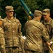 A U.S. Army military ceremony is taking place outdoors, likely a change of command or unit activation event. Several soldiers in MultiCam/OCP camouflage uniforms are gathered, with one soldier holding a teal/blue unit guidon or colors. Name tapes visible include 'LATHAM' and partially 'KIRKPATRICK', and rank insignia suggest senior officers are present.