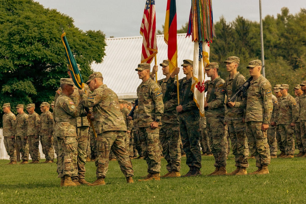 A formal U.S. Army military ceremony is taking place on a grass parade field, likely a change of command or unit activation/deactivation ceremony. Soldiers in MultiCam (OCP) uniforms are arranged in formation while a color guard holds multiple flags including the U.S. flag and what appears to be a German flag. Two senior soldiers appear to be exchanging or presenting a guidon in the foreground.