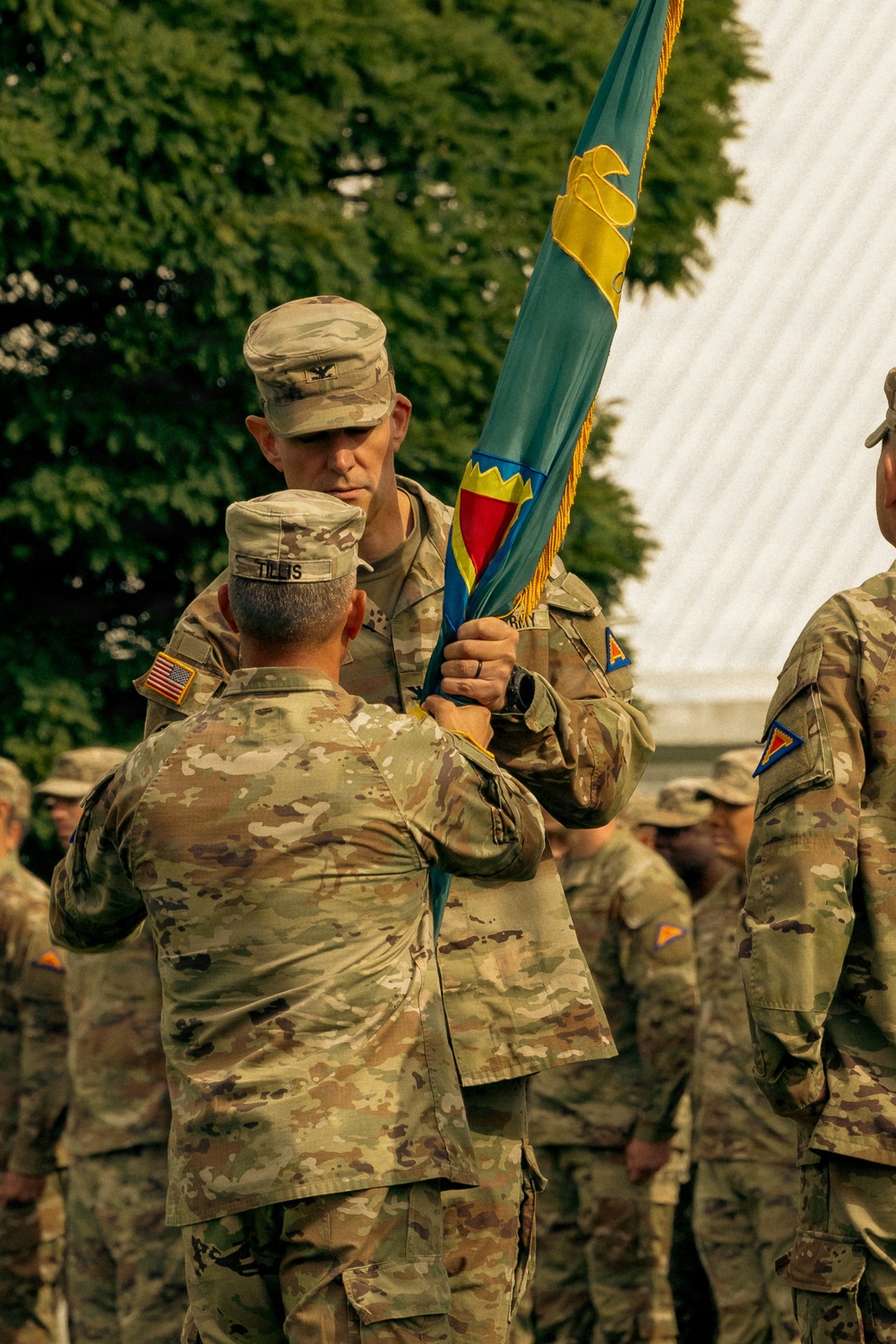 A U.S. Army change of command or activation ceremony is taking place outdoors, with two soldiers exchanging a unit guidon (flag). The soldier facing the camera wears a Colonel's rank insignia (eagle) on his patrol cap, and the soldier with his back to the camera has the name tape 'TILLIS' visible. Multiple soldiers in OCP (Operational Camouflage Pattern) uniforms are present in the background.