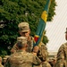 A U.S. Army change of command or activation ceremony is taking place outdoors, with two soldiers exchanging a unit guidon (flag). The soldier facing the camera wears a Colonel's rank insignia (eagle) on his patrol cap, and the soldier with his back to the camera has the name tape 'TILLIS' visible. Multiple soldiers in OCP (Operational Camouflage Pattern) uniforms are present in the background.