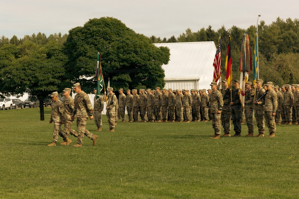 A military formation ceremony is taking place on a grass parade field, with soldiers in camouflage uniforms standing in organized ranks. Multiple flags are visible including the United States flag and the German flag, suggesting a multinational or NATO-affiliated event. A small group of soldiers appears to be marching or walking in front of the main formation, consistent with a change of command or unit review ceremony.