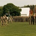 A military formation ceremony is taking place on a grass parade field, with soldiers in camouflage uniforms standing in organized ranks. Multiple flags are visible including the United States flag and the German flag, suggesting a multinational or NATO-affiliated event. A small group of soldiers appears to be marching or walking in front of the main formation, consistent with a change of command or unit review ceremony.