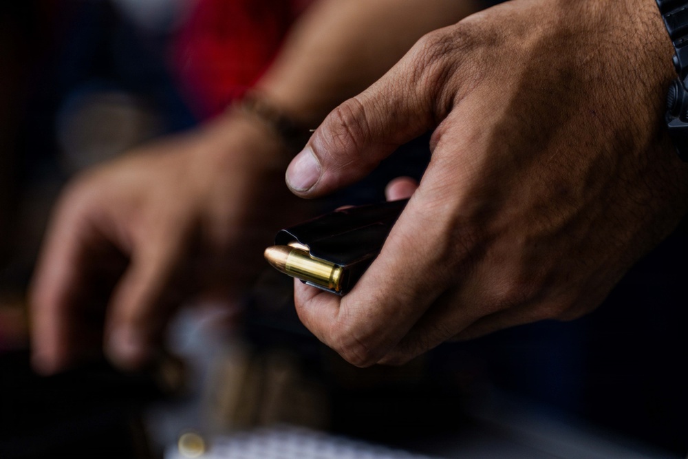 A close-up photograph shows a person's hands loading ammunition into a firearm magazine. One brass-cased cartridge is clearly visible being inserted into a dark-colored magazine. A watch is visible on the person's wrist, and a second person's hand is partially visible in the blurred background.