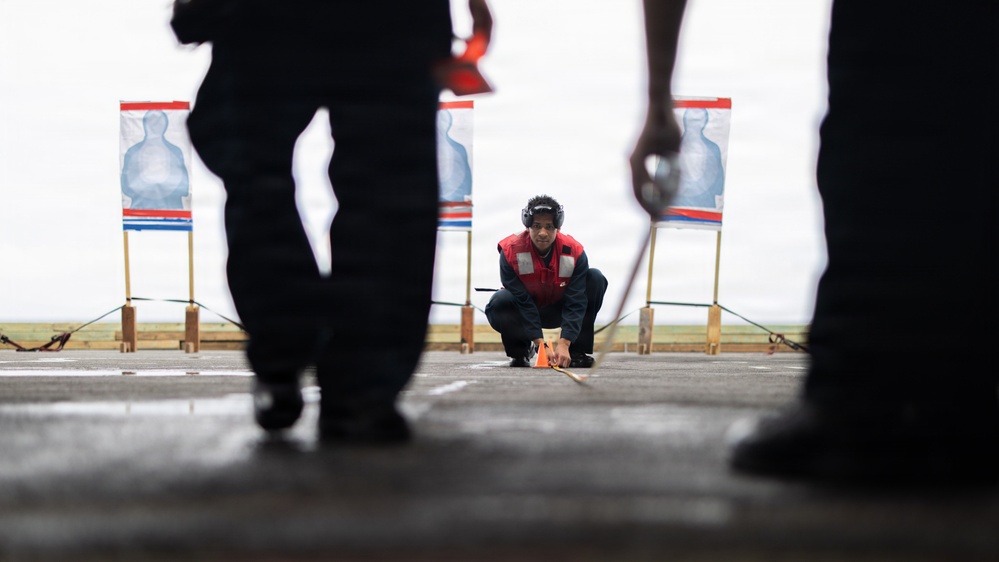 A person wearing a red vest and hearing protection is crouched down on a wet surface, appearing to place or retrieve an orange marker at what is clearly a shooting range. Three humanoid paper targets on wooden stands are visible in the background. Two silhouetted figures stand in the foreground, framing the scene from a low-angle perspective.
