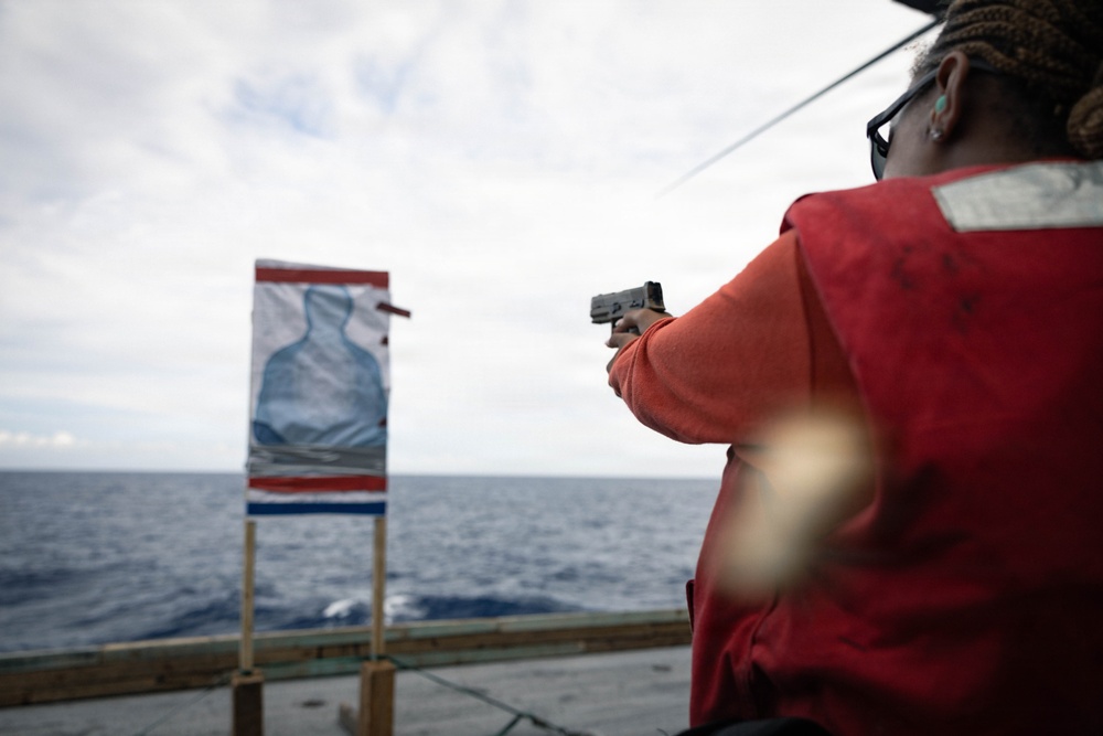 A person wearing a red vest and orange long-sleeve shirt is aiming a handgun at a silhouette target mounted on a wooden stand aboard what appears to be a ship's deck. The target features a blue human-shaped silhouette with red and blue border markings. The setting is an open ocean environment under an overcast sky.