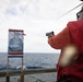 A person wearing a red vest and orange long-sleeve shirt is aiming a handgun at a silhouette target mounted on a wooden stand aboard what appears to be a ship's deck. The target features a blue human-shaped silhouette with red and blue border markings. The setting is an open ocean environment under an overcast sky.