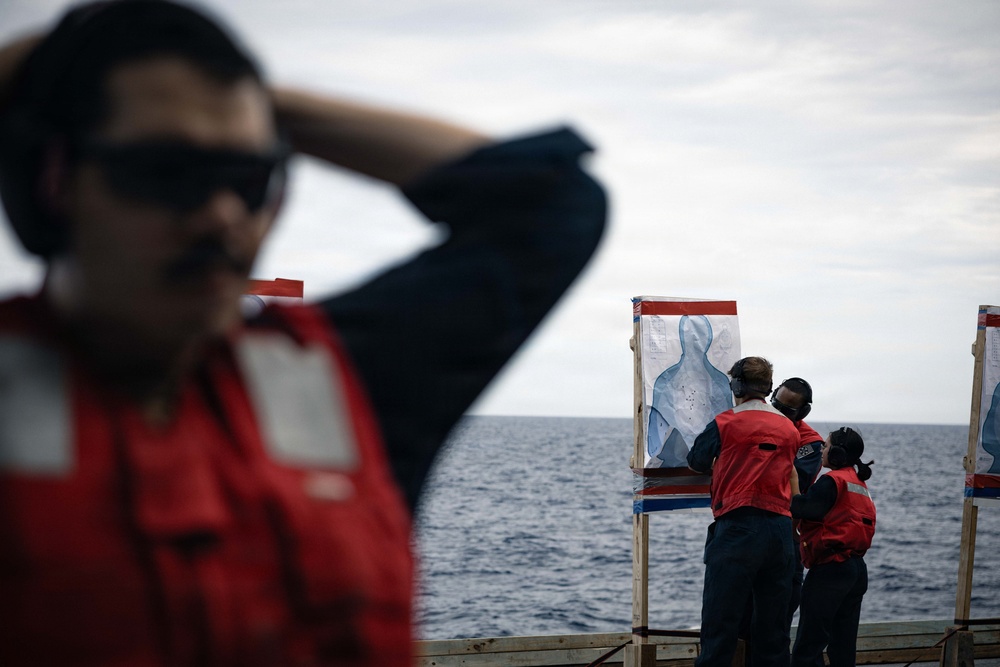 Personnel wearing red safety vests and hearing protection are examining shooting targets on what appears to be the deck of a naval vessel at sea. The targets are standard humanoid silhouette paper targets mounted on wooden frames, showing visible bullet impact marks. This appears to be a marksmanship qualification or firearms training exercise conducted aboard a ship.
