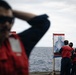 Personnel wearing red safety vests and hearing protection are examining shooting targets on what appears to be the deck of a naval vessel at sea. The targets are standard humanoid silhouette paper targets mounted on wooden frames, showing visible bullet impact marks. This appears to be a marksmanship qualification or firearms training exercise conducted aboard a ship.