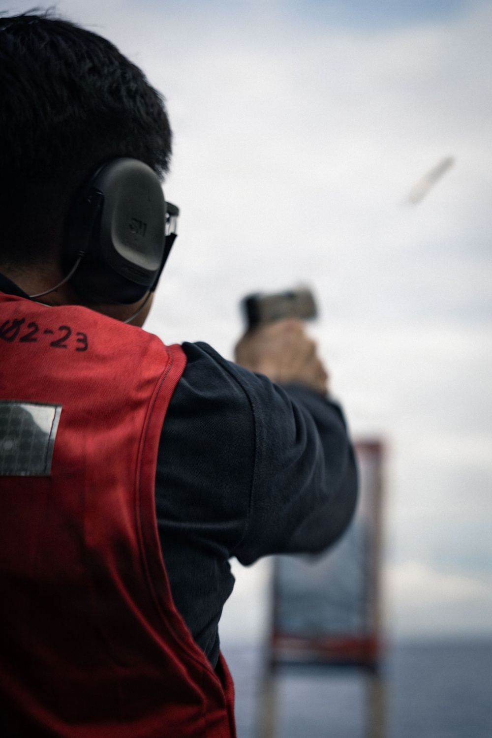 A person is firing a pistol at an outdoor shooting range, captured from a rear-angle perspective. A spent brass casing is visibly ejecting from the firearm mid-shot. The shooter is wearing hearing protection and a red vest marked with the identifier '02-23'.