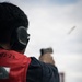 A person is firing a pistol at an outdoor shooting range, captured from a rear-angle perspective. A spent brass casing is visibly ejecting from the firearm mid-shot. The shooter is wearing hearing protection and a red vest marked with the identifier '02-23'.