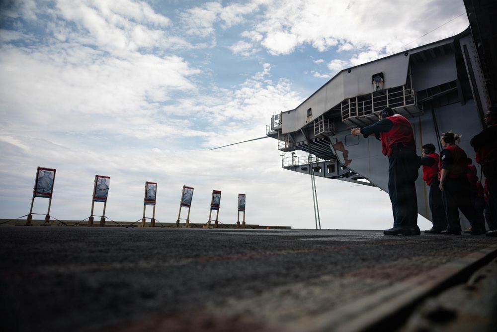 Military personnel in red jackets are conducting a firearms qualification exercise on the flight deck of a naval vessel, firing at a row of paper targets set up along the deck. The low-angle perspective emphasizes the line of targets extending toward the horizon, with the ship's island superstructure visible on the right. The scene depicts a standard shipboard small arms training evolution.