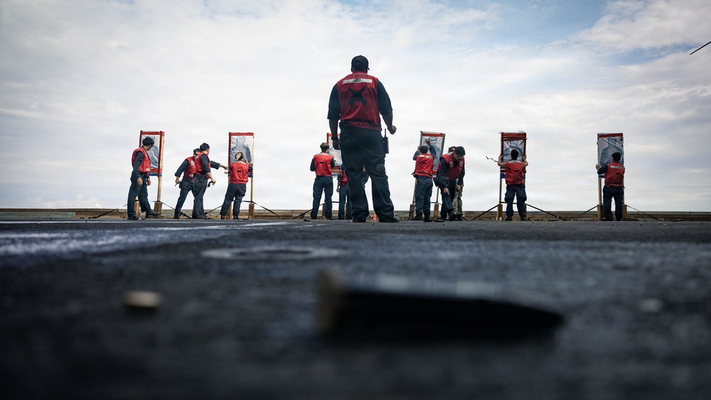 A group of personnel wearing red safety vests are engaged in a firearms qualification or training exercise on what appears to be the deck of a naval vessel. Multiple shooters are positioned at individual target stands arranged in a line, with a supervisor or range safety officer standing centrally with their back to the camera. The low-angle perspective from the deck surface emphasizes the scale and organization of the exercise.