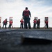 A group of personnel wearing red safety vests are engaged in a firearms qualification or training exercise on what appears to be the deck of a naval vessel. Multiple shooters are positioned at individual target stands arranged in a line, with a supervisor or range safety officer standing centrally with their back to the camera. The low-angle perspective from the deck surface emphasizes the scale and organization of the exercise.