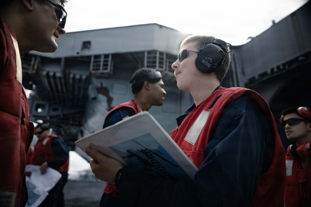 Several personnel wearing red safety vests and hearing protection are gathered on what appears to be a naval vessel deck. A central figure holds a document or booklet and appears to be in conversation with another crew member. The background shows the superstructure of a large warship, likely an aircraft carrier or amphibious assault ship.
