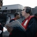 Several personnel wearing red safety vests and hearing protection are gathered on what appears to be a naval vessel deck. A central figure holds a document or booklet and appears to be in conversation with another crew member. The background shows the superstructure of a large warship, likely an aircraft carrier or amphibious assault ship.