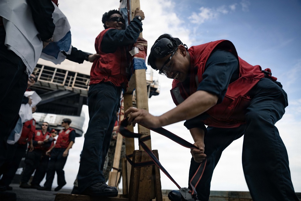 Several personnel wearing red safety vests and dark uniforms are working on what appears to be a naval vessel deck. The foreground figure is bent over handling straps or lines secured to a wooden post structure, while others assist nearby. The scene suggests a ceremonial or operational deck activity aboard a military ship.