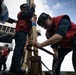Several personnel wearing red safety vests and dark uniforms are working on what appears to be a naval vessel deck. The foreground figure is bent over handling straps or lines secured to a wooden post structure, while others assist nearby. The scene suggests a ceremonial or operational deck activity aboard a military ship.