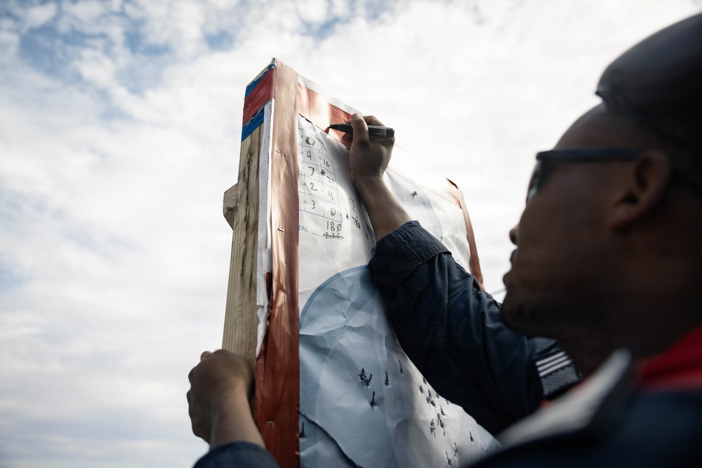 A person wearing glasses and a dark jacket is writing on a paper scorecard or tally sheet attached to a wooden post. The sheet appears to contain rows of numbers, suggesting score-keeping or tallying activity. The photo is taken from a low angle against an overcast sky.