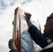 A person wearing glasses and a dark jacket is writing on a paper scorecard or tally sheet attached to a wooden post. The sheet appears to contain rows of numbers, suggesting score-keeping or tallying activity. The photo is taken from a low angle against an overcast sky.