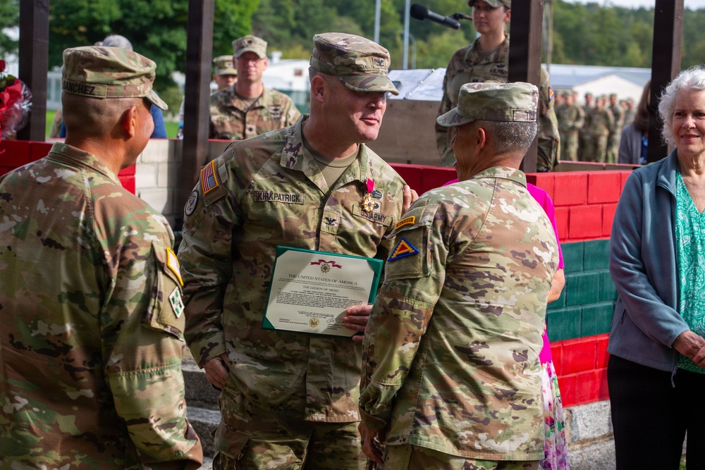 A U.S. Army military ceremony is taking place outdoors, where a soldier identified by nametape as 'KIRKPATRICK' is receiving the Legion of Merit award. He is holding a certificate reading 'The United States of America - The Legion of Merit' and wearing the medal on his uniform. Several other uniformed soldiers and civilians are present, suggesting a formal awards or retirement ceremony.