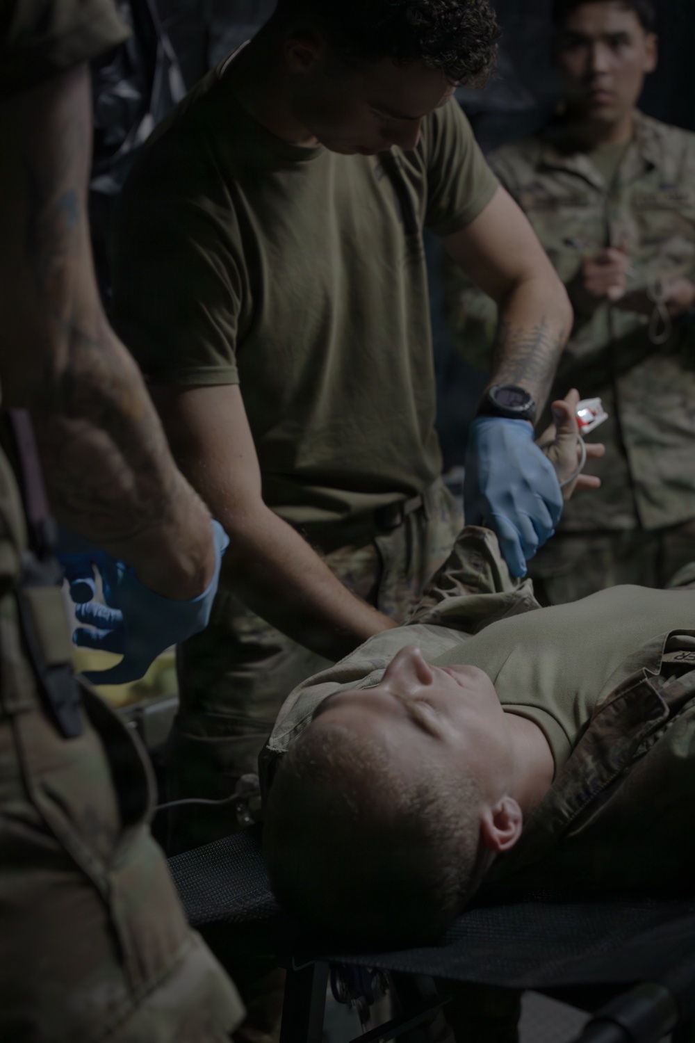 Military personnel in olive drab uniforms are performing what appears to be a tactical combat casualty care (TCCC) scenario or medical training exercise. A provider wearing blue nitrile gloves is attending to a supine casualty on a litter or stretcher. A second uniformed individual observes in the background.