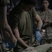 Military personnel in olive drab uniforms are performing what appears to be a tactical combat casualty care (TCCC) scenario or medical training exercise. A provider wearing blue nitrile gloves is attending to a supine casualty on a litter or stretcher. A second uniformed individual observes in the background.