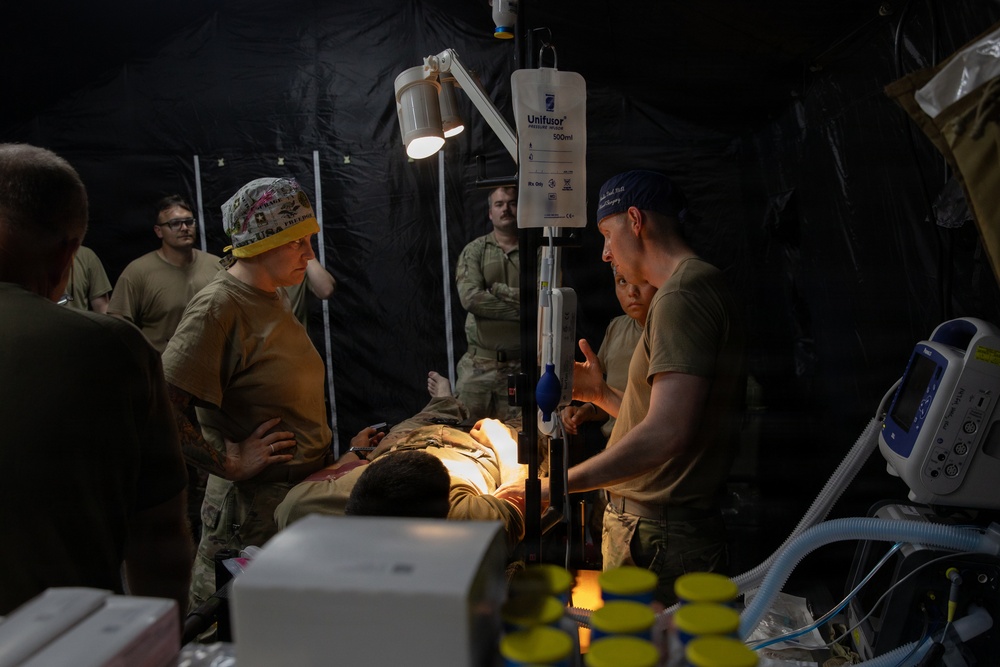 Military medical personnel are treating a patient on a field surgical table inside a dark tactical tent or field hospital. Medical staff wearing scrub caps and military uniforms are gathered around the patient, with IV equipment and monitoring devices visible. The scene depicts a forward surgical or trauma care environment, likely during a military exercise or real-world operation.