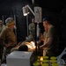 Military medical personnel are treating a patient on a field surgical table inside a dark tactical tent or field hospital. Medical staff wearing scrub caps and military uniforms are gathered around the patient, with IV equipment and monitoring devices visible. The scene depicts a forward surgical or trauma care environment, likely during a military exercise or real-world operation.
