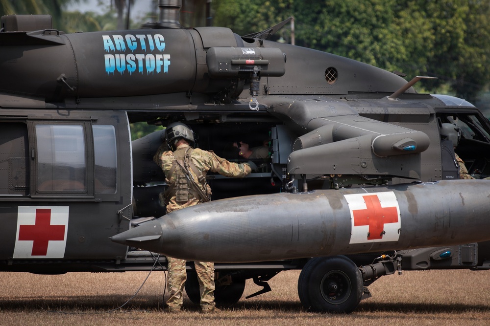 A military medical evacuation (MEDEVAC) helicopter, specifically a UH-60 Black Hawk, is shown on the ground with the callsign 'Arctic Dustoff' painted on its fuselage. The aircraft bears Red Cross markings on both sides, identifying it as a medical helicopter. At least two personnel in military camouflage uniforms and flight helmets are visible, with one appearing to conduct pre-flight checks or maintenance at the cabin door.