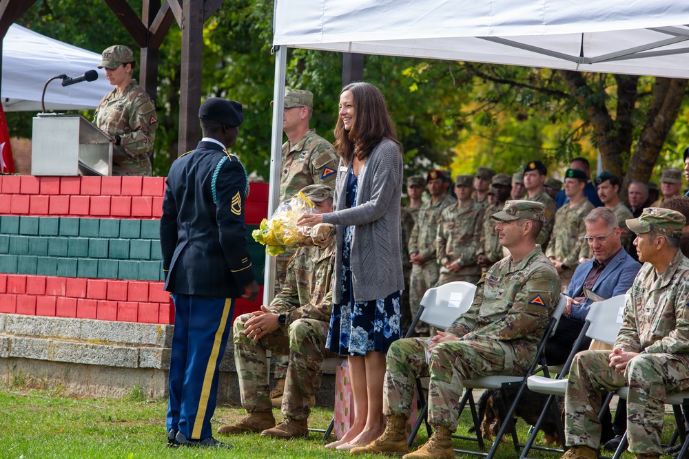 A formal U.S. Army military ceremony is taking place outdoors under a white canopy tent. A soldier in dress blues is presenting yellow flowers to a civilian woman who is smiling, while rows of uniformed soldiers and a few civilians observe from seated positions. A female soldier is visible at a podium with a microphone in the background.
