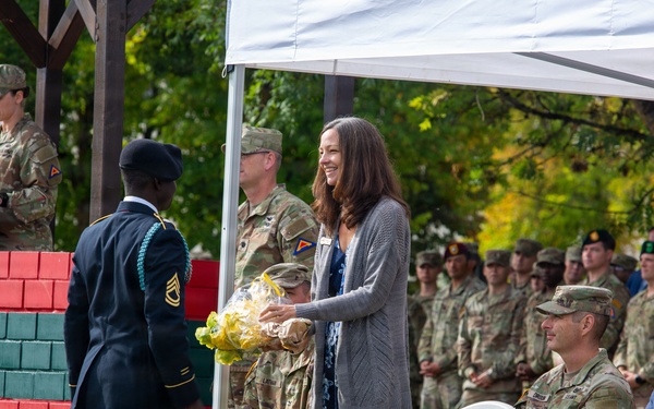A formal U.S. Army military ceremony is taking place outdoors under a white canopy tent. A soldier in dress blues is presenting yellow flowers to a civilian woman who is smiling, while rows of uniformed soldiers and a few civilians observe from seated positions. A female soldier is visible at a podium with a microphone in the background.
