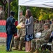 A formal U.S. Army military ceremony is taking place outdoors under a white canopy tent. A soldier in dress blues is presenting yellow flowers to a civilian woman who is smiling, while rows of uniformed soldiers and a few civilians observe from seated positions. A female soldier is visible at a podium with a microphone in the background.