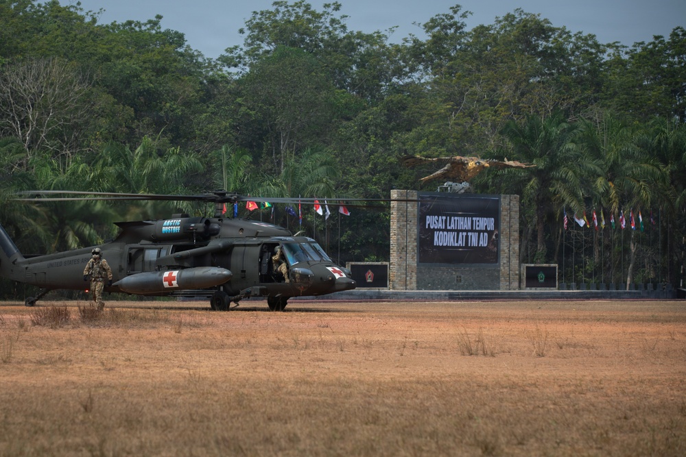 Two U.S. Army UH-60 Black Hawk helicopters marked with Red Cross medical insignia are parked on a dry grass field at the Pusat Latihan Tempur Kodiklat TNI AD (Indonesian Army Combat Training Center). At least two personnel in military gear are visible near the aircraft. Multiple national flags are displayed at the facility entrance, indicating a multinational military exercise.