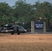 Two U.S. Army UH-60 Black Hawk helicopters marked with Red Cross medical insignia are parked on a dry grass field at the Pusat Latihan Tempur Kodiklat TNI AD (Indonesian Army Combat Training Center). At least two personnel in military gear are visible near the aircraft. Multiple national flags are displayed at the facility entrance, indicating a multinational military exercise.