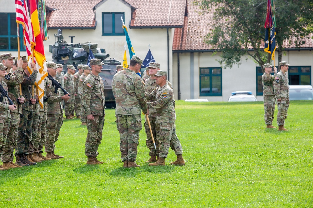 A U.S. Army change of command or activation ceremony is taking place on a grassy parade field, likely at a military installation in Europe based on the architecture. Two senior soldiers are exchanging a unit guidon (flag) in the center, which is the traditional symbolic act of transferring command authority. Formations of soldiers in OCP (Operational Camouflage Pattern) uniforms flank the ceremony, with color guards bearing multiple flags including the U.S. flag.