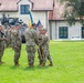 A U.S. Army change of command or activation ceremony is taking place on a grassy parade field, likely at a military installation in Europe based on the architecture. Two senior soldiers are exchanging a unit guidon (flag) in the center, which is the traditional symbolic act of transferring command authority. Formations of soldiers in OCP (Operational Camouflage Pattern) uniforms flank the ceremony, with color guards bearing multiple flags including the U.S. flag.