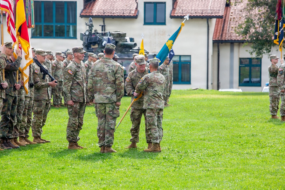 A U.S. Army military ceremony, likely a change of command or activation ceremony, is taking place on a grassy parade field. Three central figures are engaged in the passing of a unit guidon (flag), which is the symbolic focal point of the event. Soldiers in Army Combat Uniforms (ACU/OCP pattern) are formed in ranks on both sides, with multiple flags and guidons visible.
