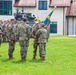 A U.S. Army military ceremony, likely a change of command or activation ceremony, is taking place on a grassy parade field. Three central figures are engaged in the passing of a unit guidon (flag), which is the symbolic focal point of the event. Soldiers in Army Combat Uniforms (ACU/OCP pattern) are formed in ranks on both sides, with multiple flags and guidons visible.